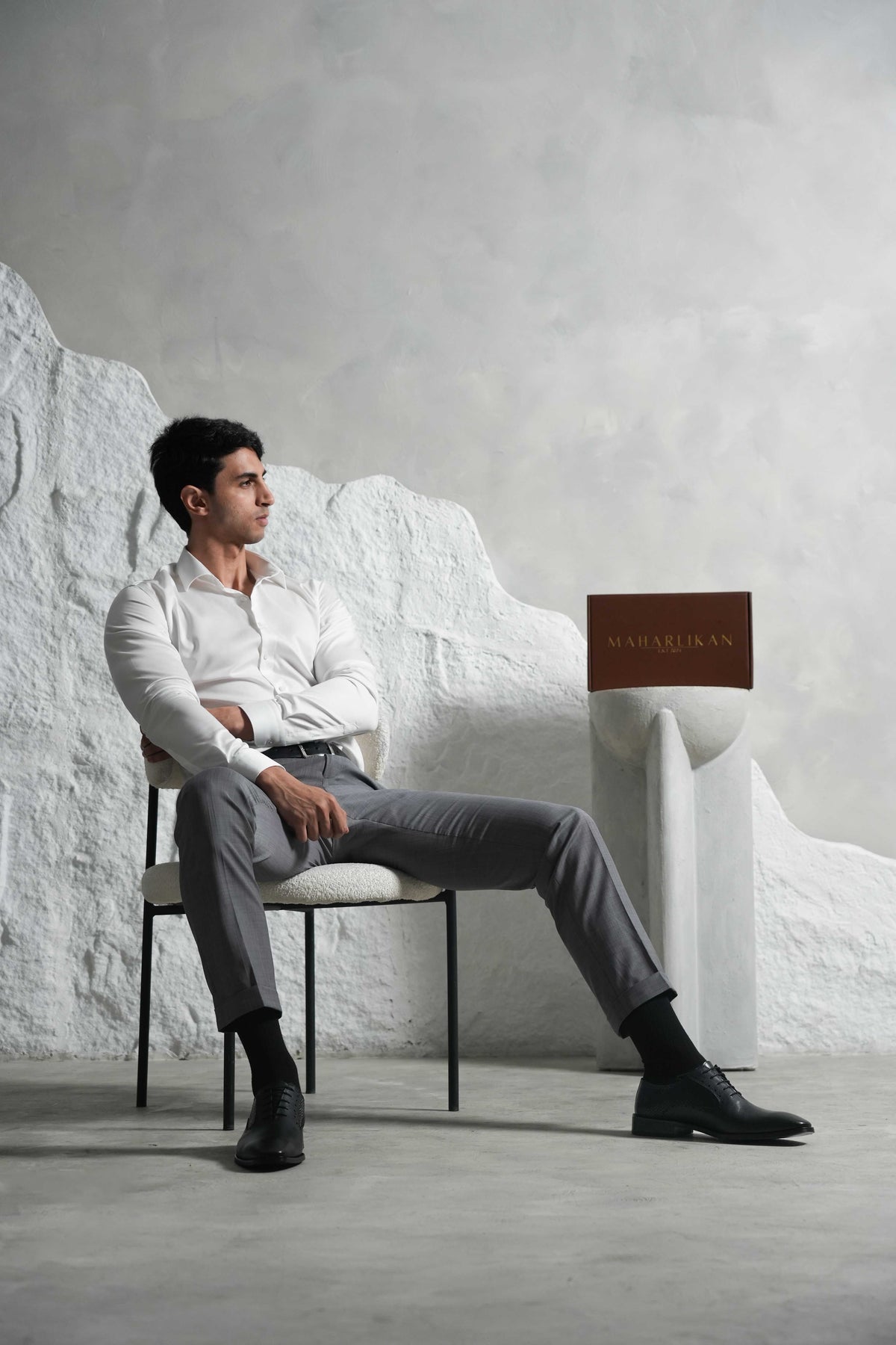 A stylish man sits confidently in a modern setting wearing a white shirt, gray trousers, and black shoes. Beside him is a pedestal with a MAHARLIKAN box, showcasing a refined, minimalist aesthetic perfect for a luxury fashion brand.